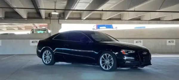 Black Audi sedan parked in a concrete multi‑level parking garage with numbered spaces in the background, glossy black finish and large alloy wheels visible.