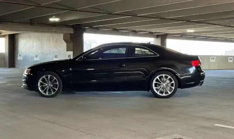 Black two-door coupe parked in a concrete parking garage, side view showing alloy wheels and tinted windows.