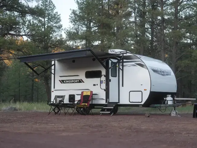 White Kingsport travel trailer parked on dirt in a pine forest, with an extended awning and two camping chairs outside.