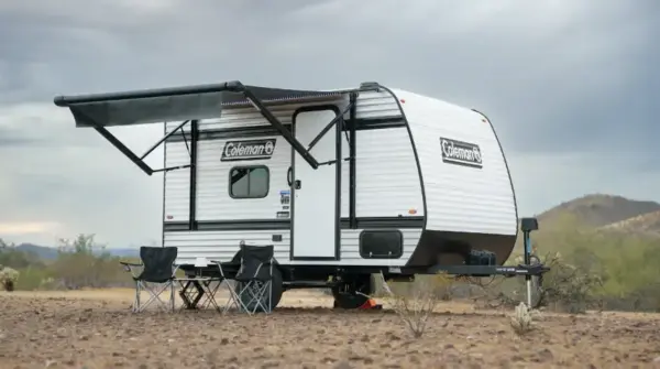 White Coleman travel trailer with awning extended, set up for camping with four black folding chairs on a dusty desert site.