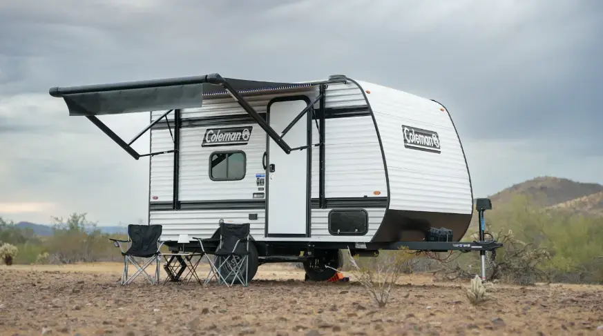 White Coleman travel trailer with awning extended, set up for camping with four black folding chairs on a dusty desert site.