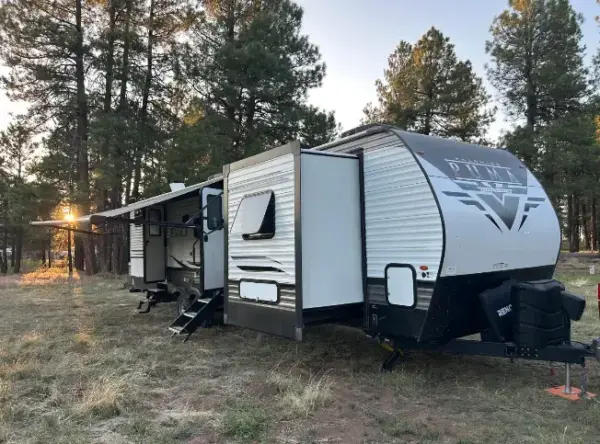 Large modern travel trailer with slide-outs parked on a grassy forest site at dusk, awning extended and steps down by the entrance in a pine-filled area.