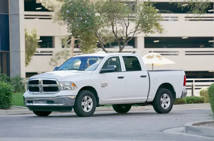 White pickup truck parked in a parking lot with a multi-story parking structure and trees nearby.